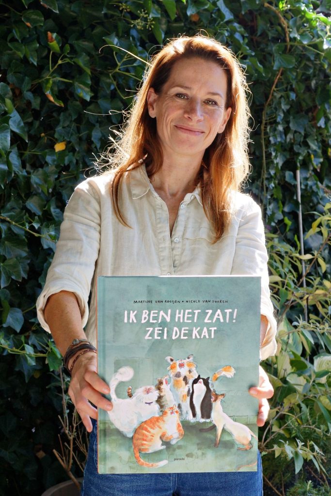 Portret van een maker die lachend een prentenboek vasthoudt, voor een muur met klimop