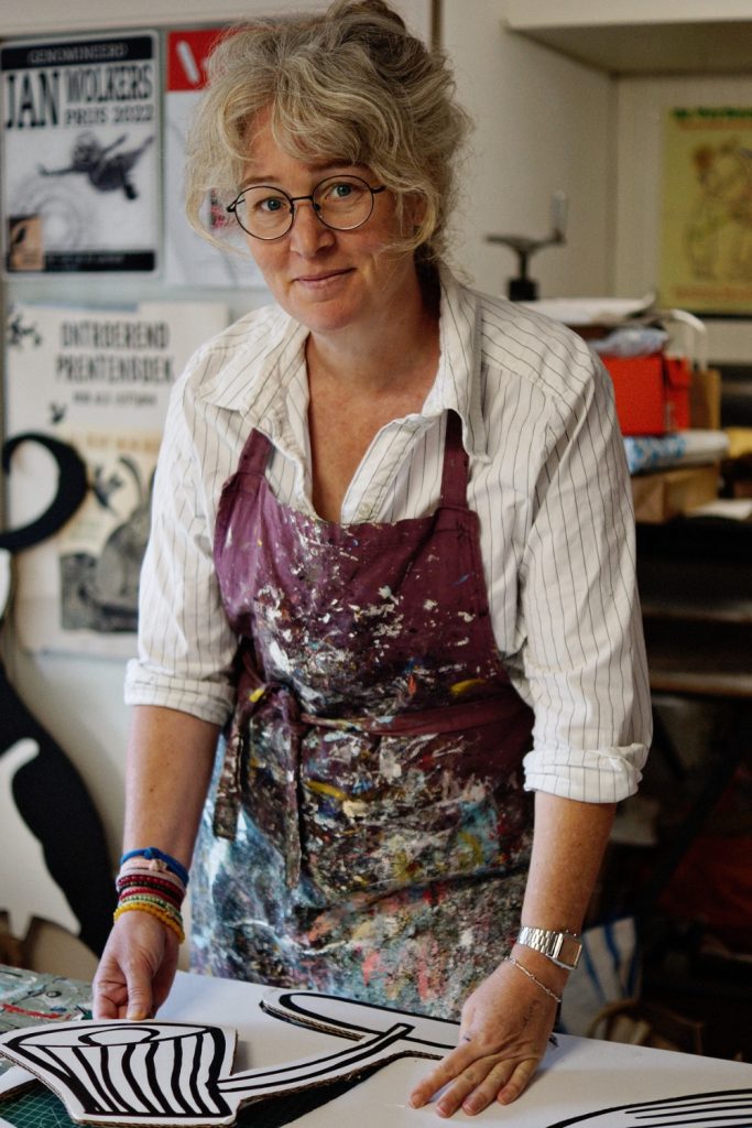 Portret van een maker in een met verf bespatte schort aan een werktafel met grote kartonnen uitsnedes in het atelier